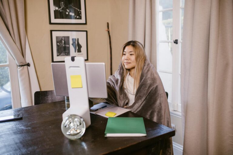 A woman wrapped in a blanket attending an online class at home using a computer.