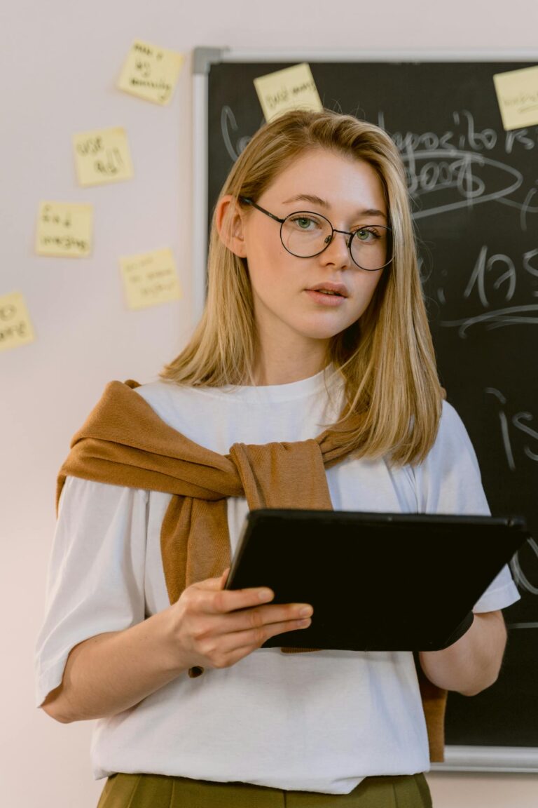 Caucasian woman standing in a classroom holding a tablet with sticky notes on a blackboard behind her.