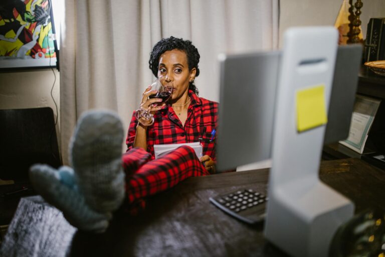Woman in red pajamas relaxing with wine at a home office desk with feet up.
